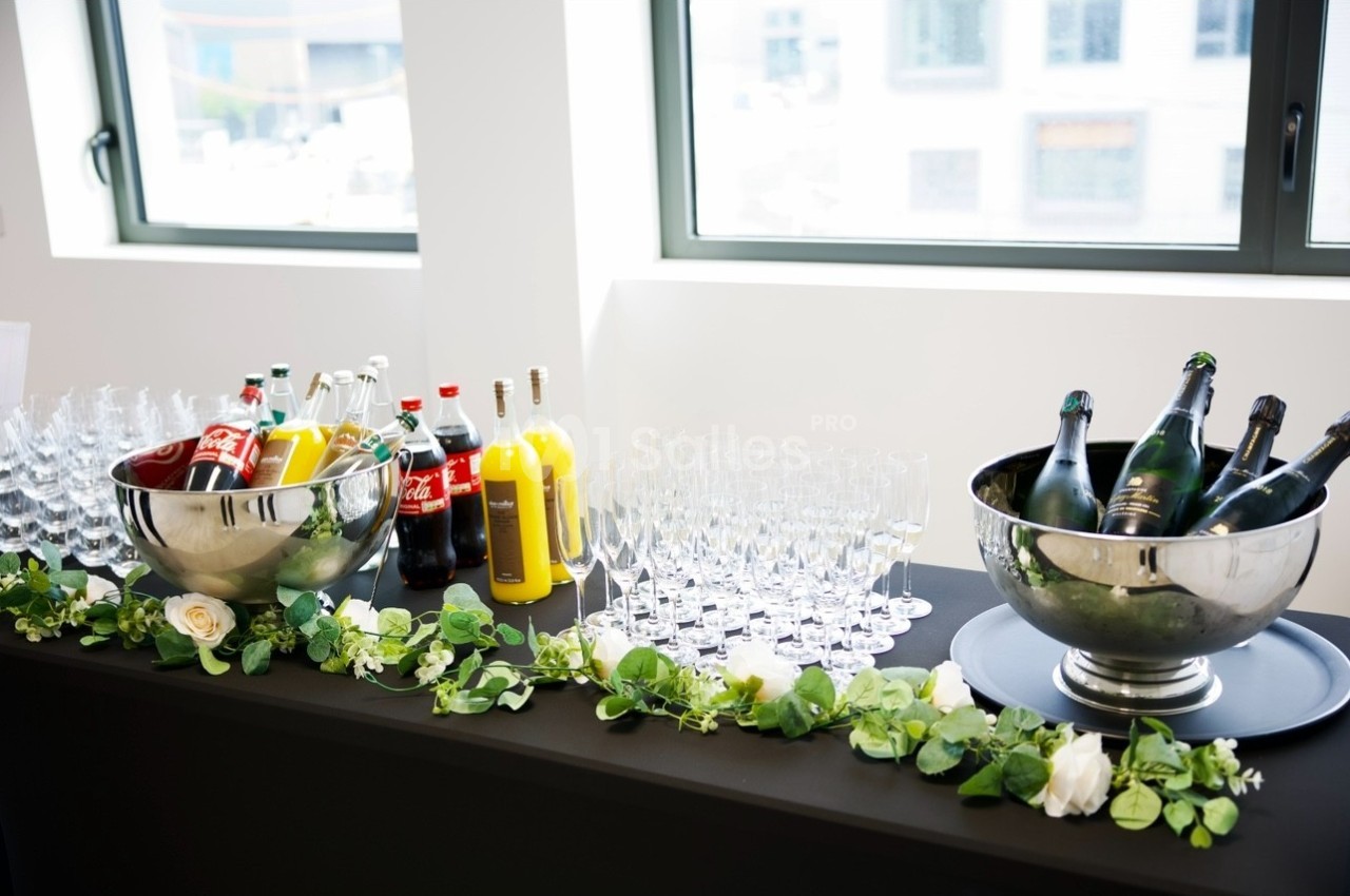 Table avec boissons variées, bouteilles de soda, jus et champagne dans des seaux, entourée de verres et décorée de verdure.