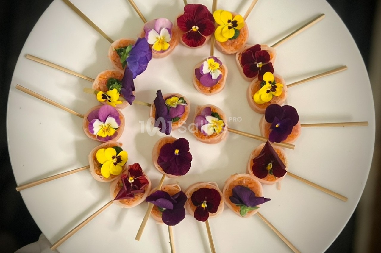 Assortiment de bouchées décorées de fleurs comestibles colorées, disposées en cercle sur une assiette blanche.