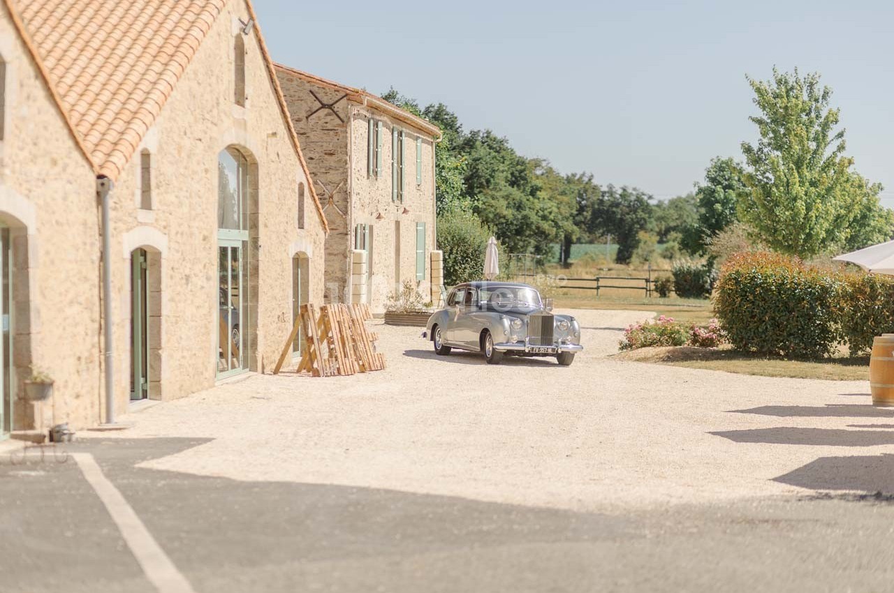 Voiture ancienne argentée garée devant des bâtiments en pierre dans une cour ensoleillée avec un jardin en arrière-plan.