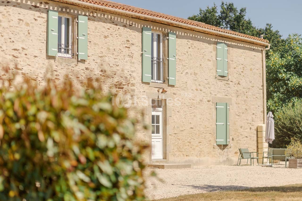 Façade en pierre d'une maison avec volets verts, entourée de végétation et d'un espace extérieur aménagé.
