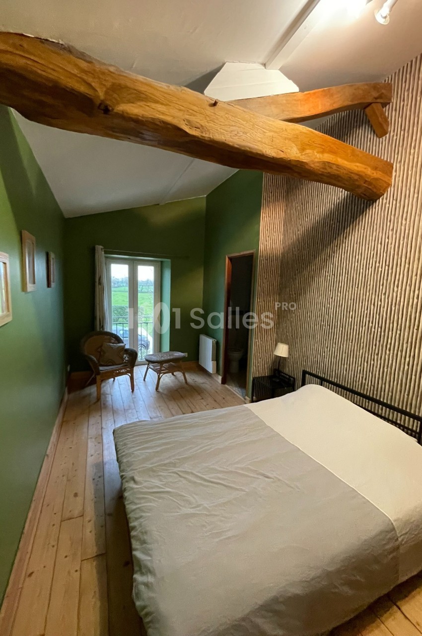 Chambre avec murs verts, lit double, parquet en bois, poutre apparente et vue sur un paysage extérieur par une porte-fenêtre.