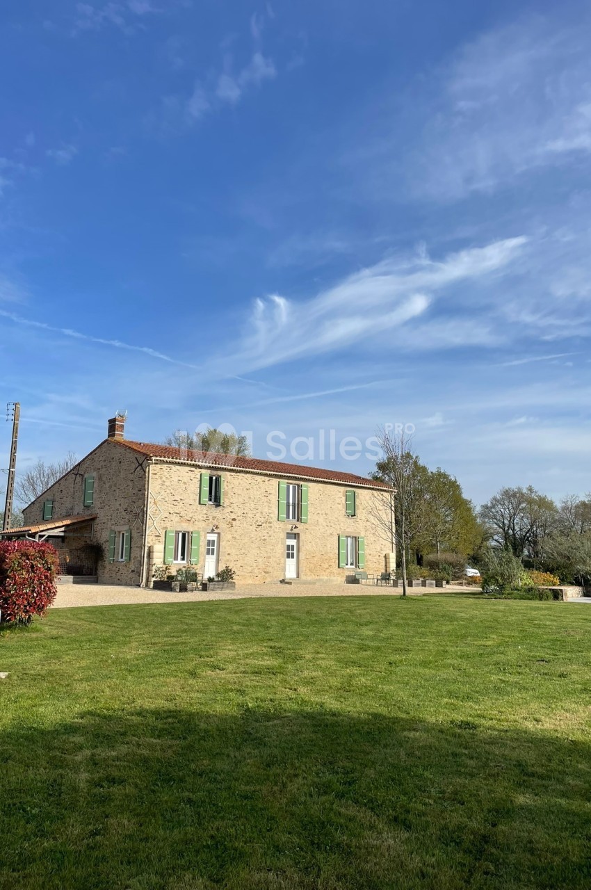Grande maison en pierre avec volets verts, entourée d'un jardin verdoyant sous un ciel bleu dégagé.