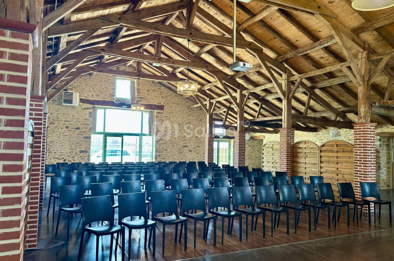 Salle avec charpente en bois, murs en pierre et rangées de chaises noires disposées pour un événement.