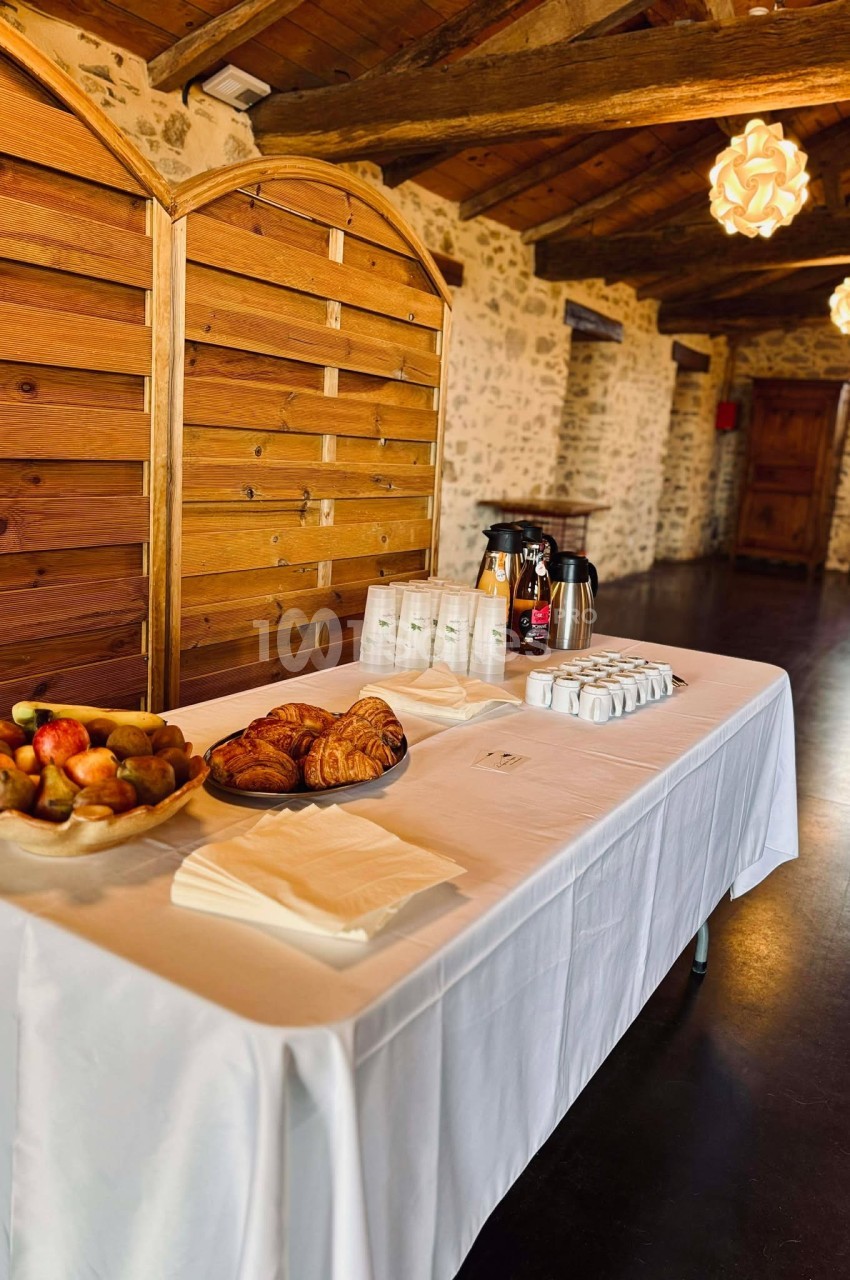 Table dressée avec viennoiseries, fruits, boissons chaudes et tasses, dans une salle en pierre et bois au plafond rustique.