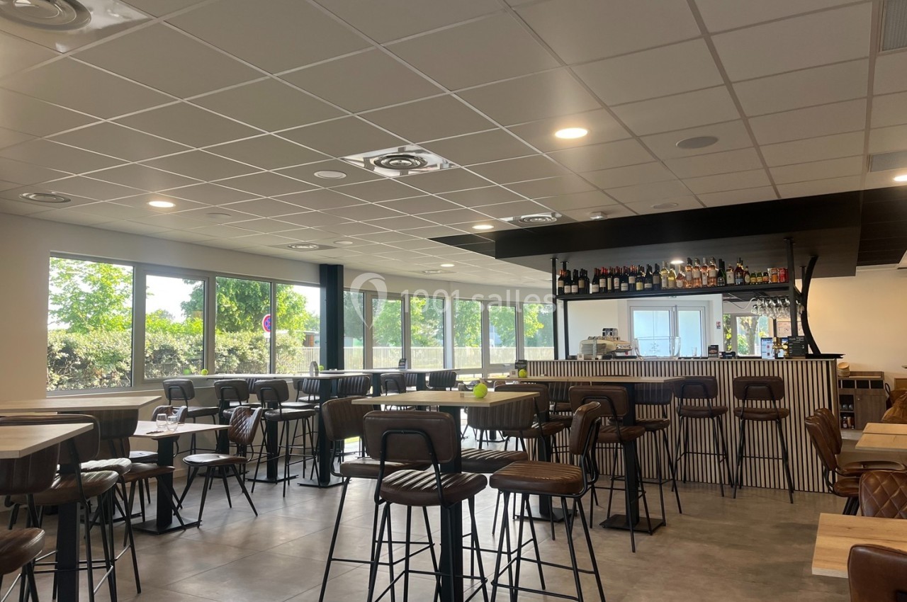 Salle lumineuse avec bar moderne, tables hautes et chaises en cuir, grandes fenêtres donnant sur un espace arboré.