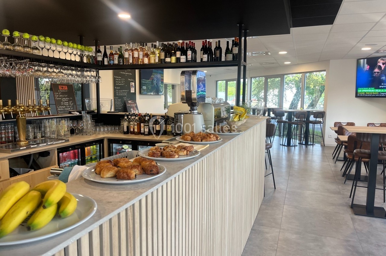 Comptoir d'un café avec viennoiseries, fruits et boissons, entouré de tables et baigné de lumière naturelle.