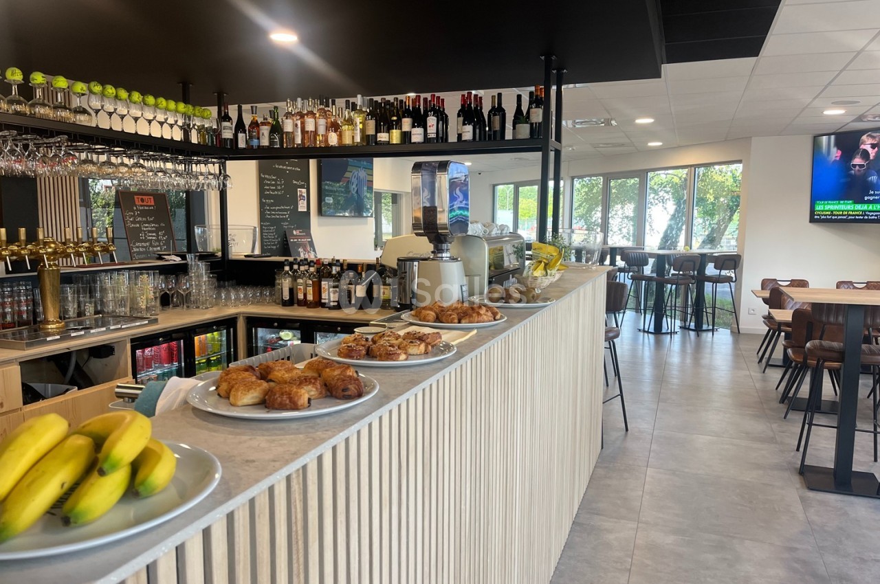 Comptoir d'un café avec viennoiseries, fruits et boissons, entouré de tables et chaises dans un espace lumineux.