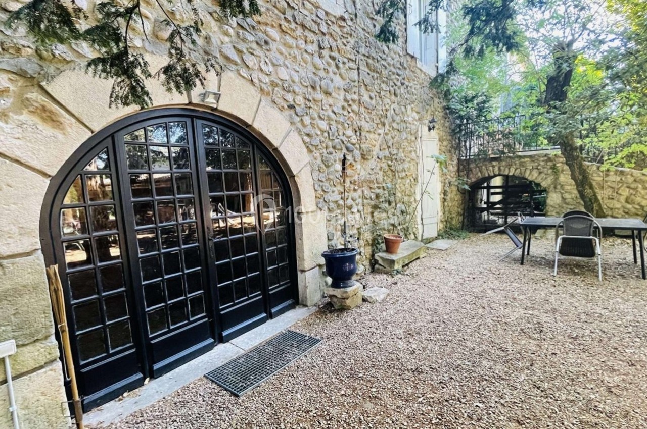 Cour extérieure avec sol en gravier, table et chaises près d'une façade en pierre avec de grandes portes vitrées cintrées.