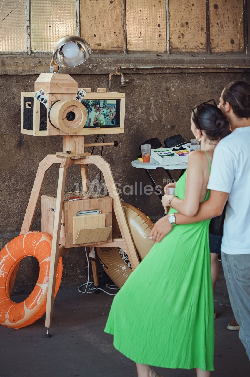 Un couple observe un photobooth en bois avec un écran intégré dans un espace industriel.
