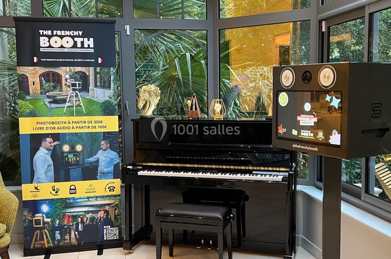 Piano noir devant une baie vitrée, accompagné d'une borne photo et d'un panneau publicitaire coloré.