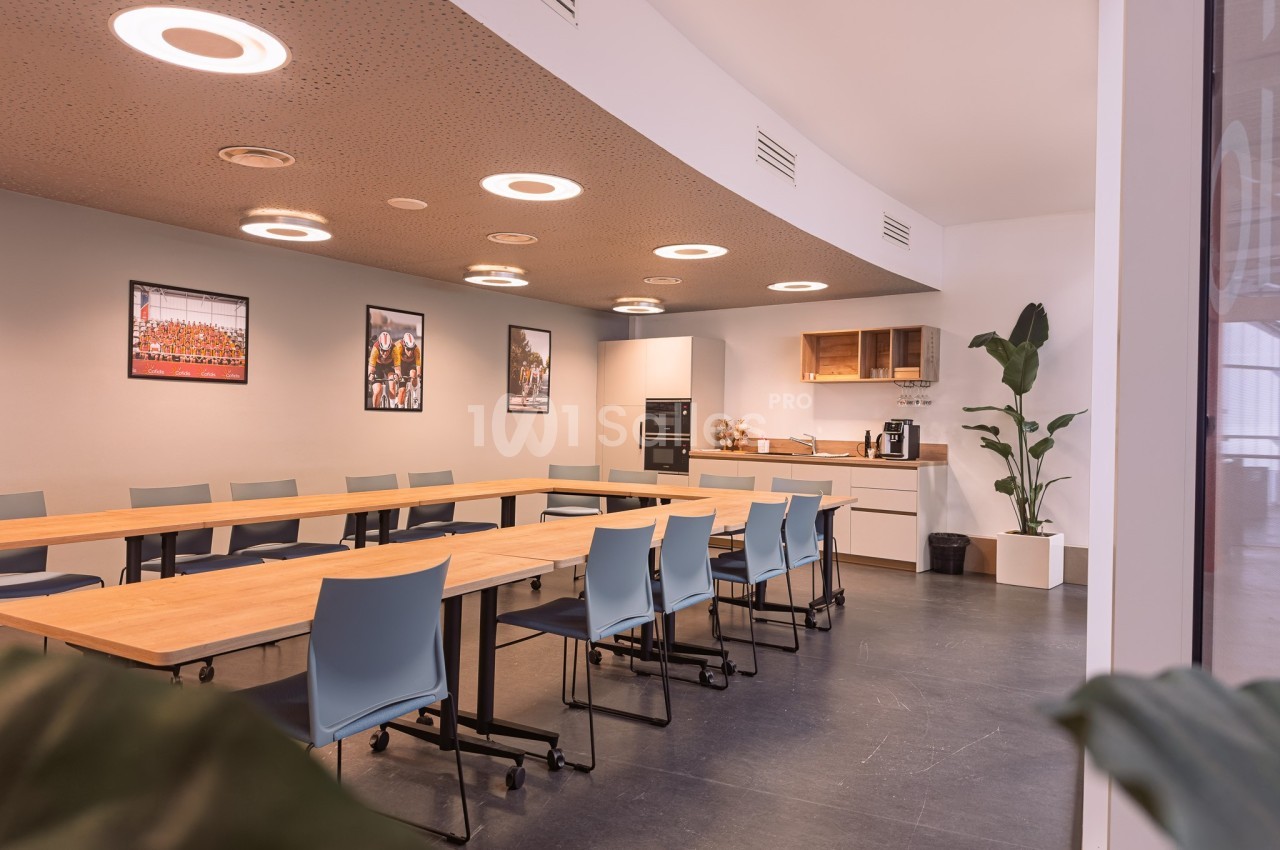 Salle de réunion lumineuse avec tables en bois, chaises bleues, kitchenette équipée et plantes décoratives.
