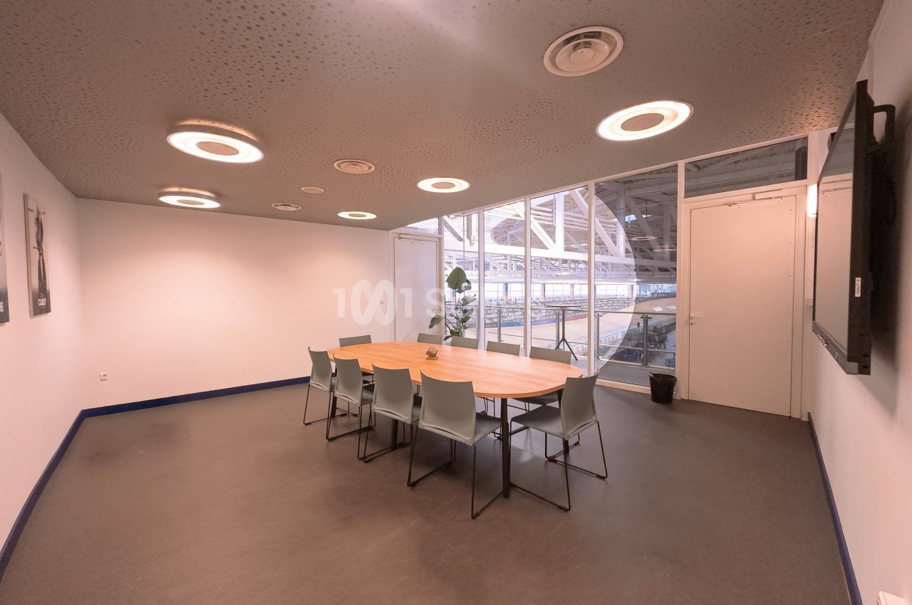 Salle de réunion lumineuse avec une table ovale en bois, des chaises grises et une vue sur un espace vitré.