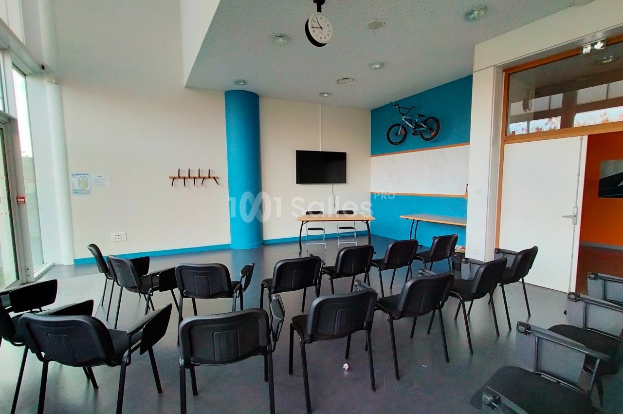 Salle de réunion avec des chaises noires disposées en rangées, un écran mural et un vélo accroché au mur.