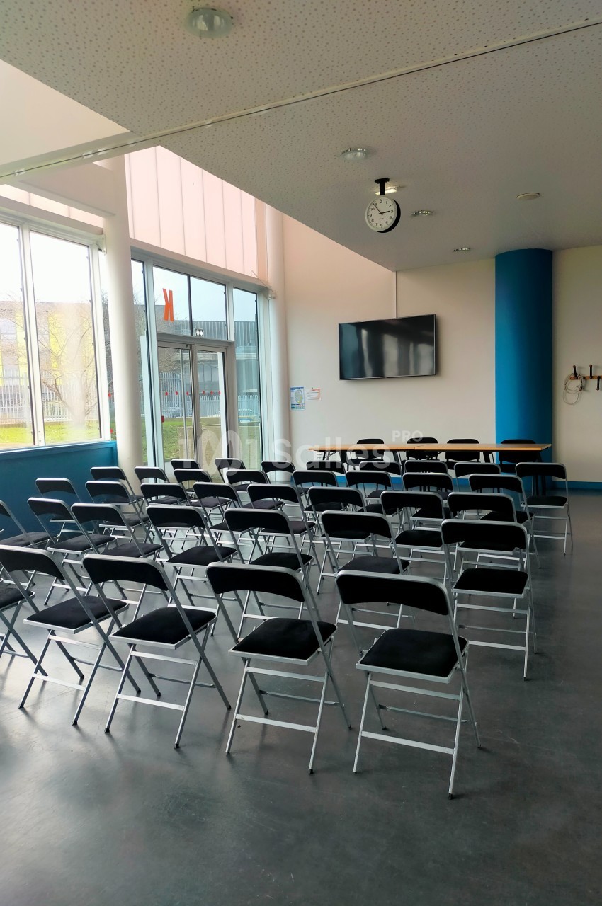 Salle de réunion lumineuse avec des rangées de chaises pliantes noires, une table, un écran mural et une horloge.