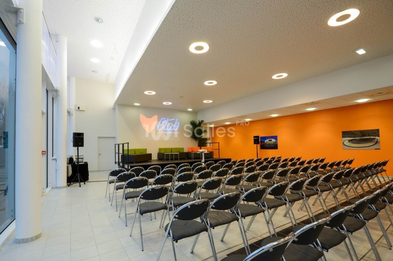 Salle de conférence lumineuse avec rangées de chaises noires, scène équipée et écran de projection allumé.