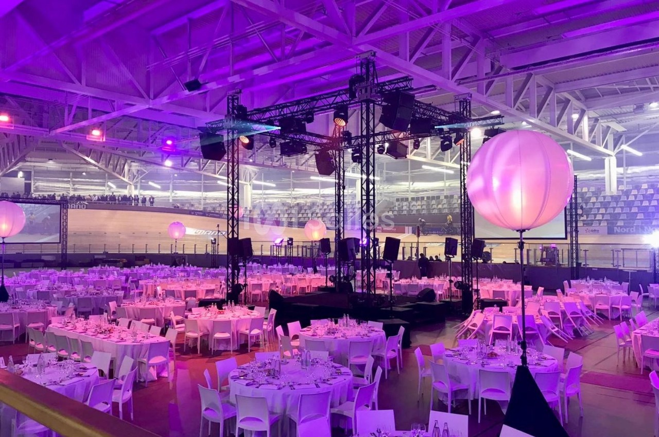 Salle de réception avec tables dressées et éclairage coloré, entourée de structures métalliques et de ballons lumineux.