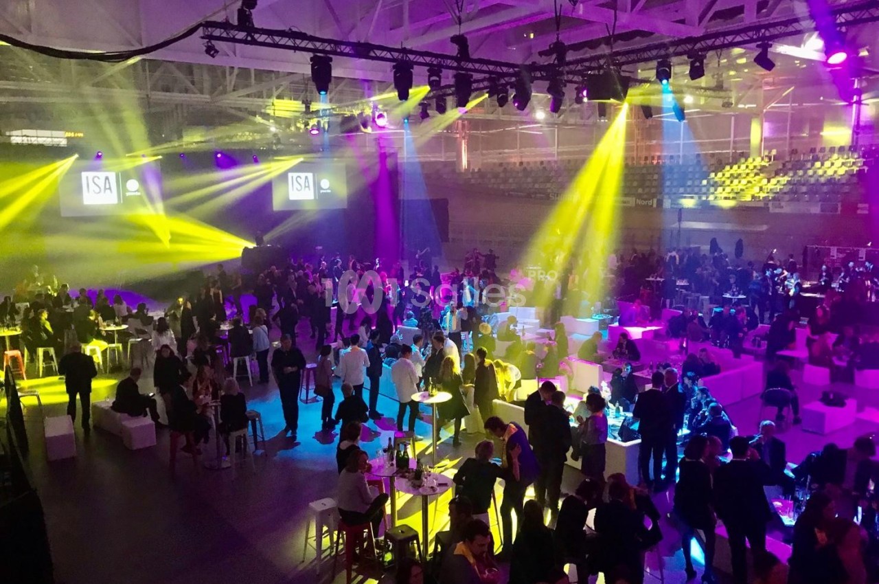 Salle de réception animée avec des invités debout ou assis, éclairée par des projecteurs colorés.