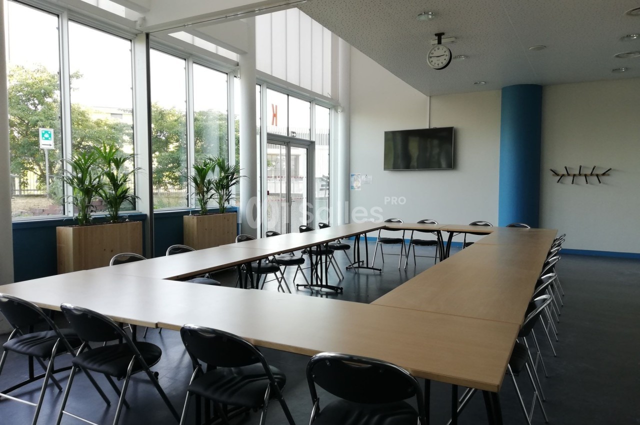 Salle de réunion lumineuse avec une grande table en U, des chaises noires, des plantes et une horloge murale.