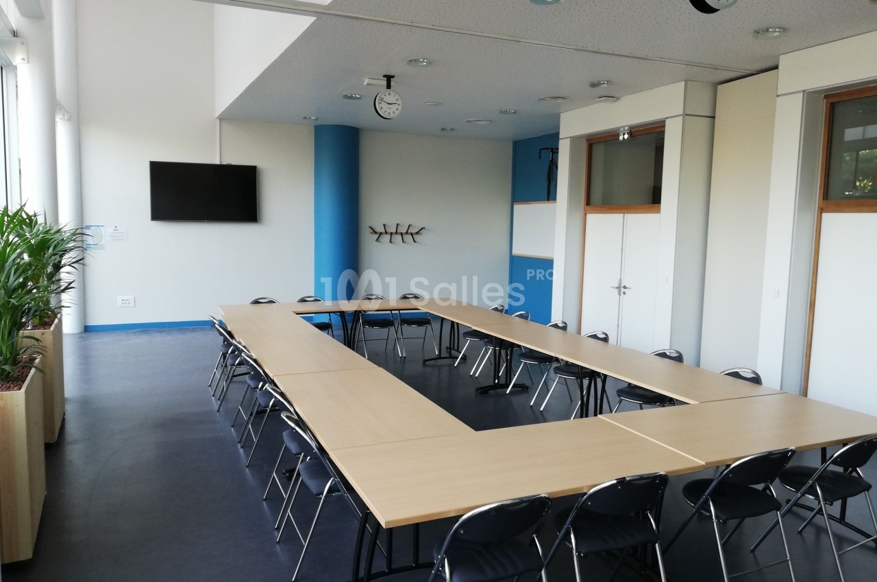 Salle de réunion lumineuse avec tables disposées en U, chaises noires, écran mural et horloges au plafond.