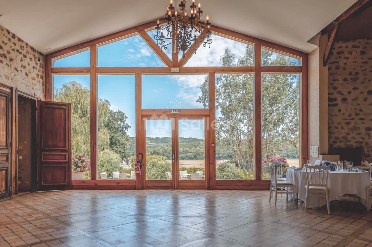 Grande salle lumineuse avec baie vitrée donnant sur un paysage verdoyant, tables dressées sur la droite.