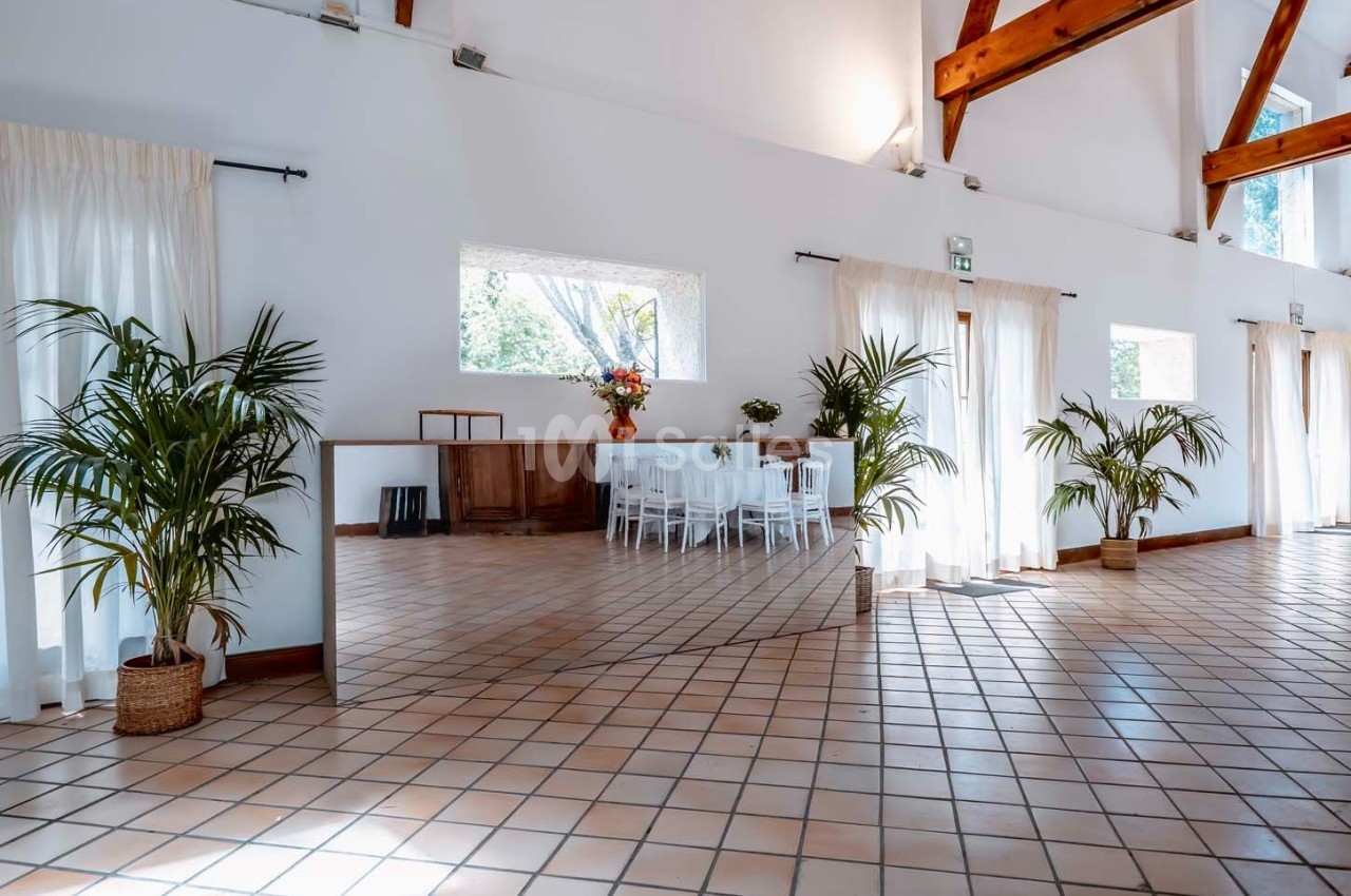 Salle lumineuse avec carrelage au sol, grandes fenêtres, plantes en pot et tables blanches dressées au fond.
