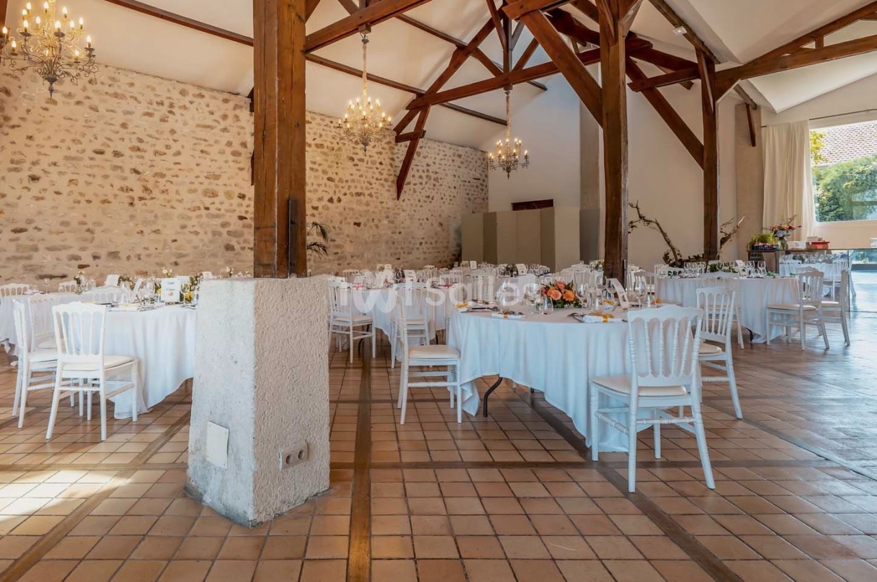 Salle de réception lumineuse avec murs en pierre, poutres apparentes, tables rondes dressées et chaises blanches.