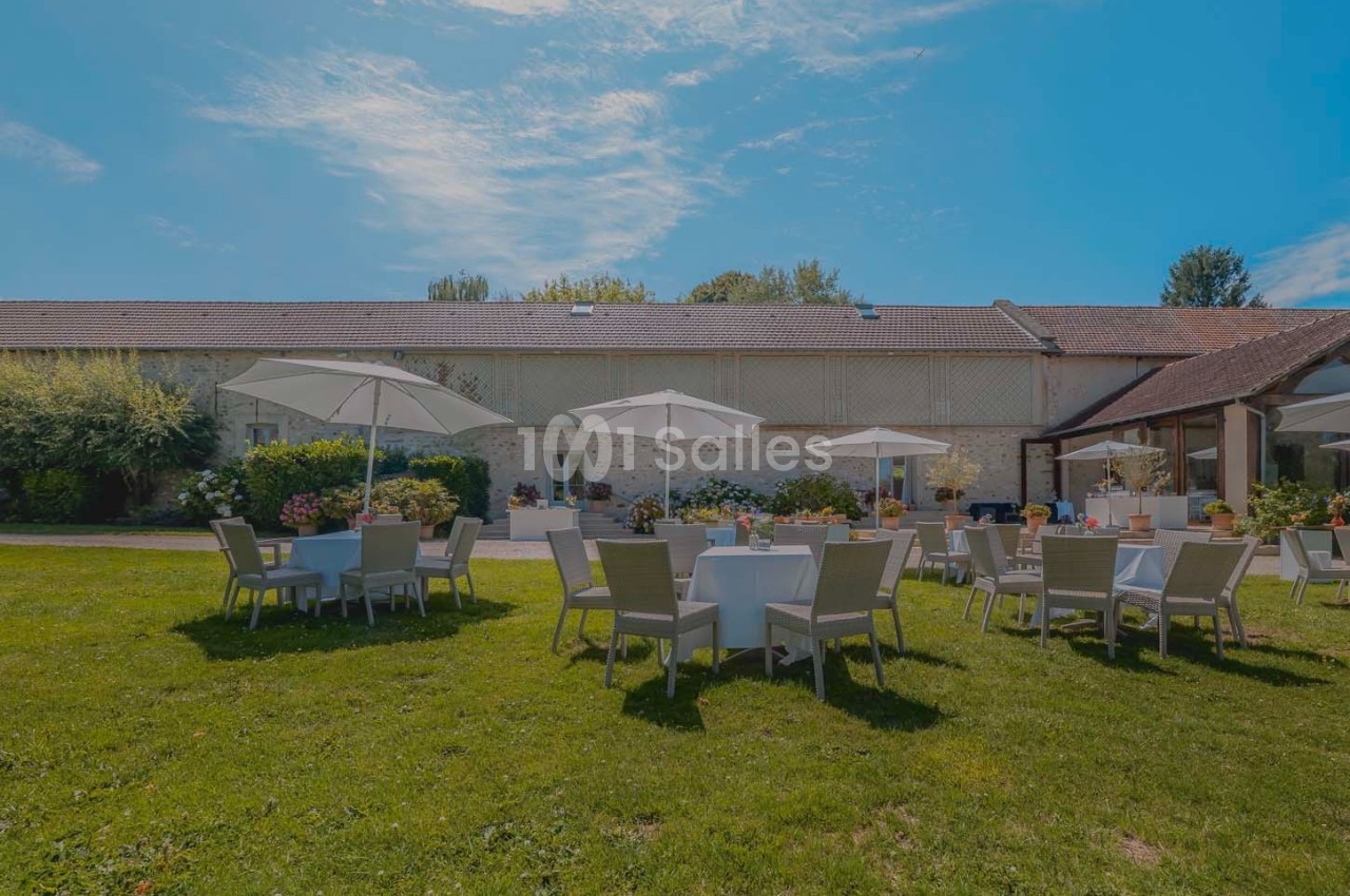 Tables et chaises disposées sur une pelouse avec parasols, devant un bâtiment en pierre sous un ciel dégagé.