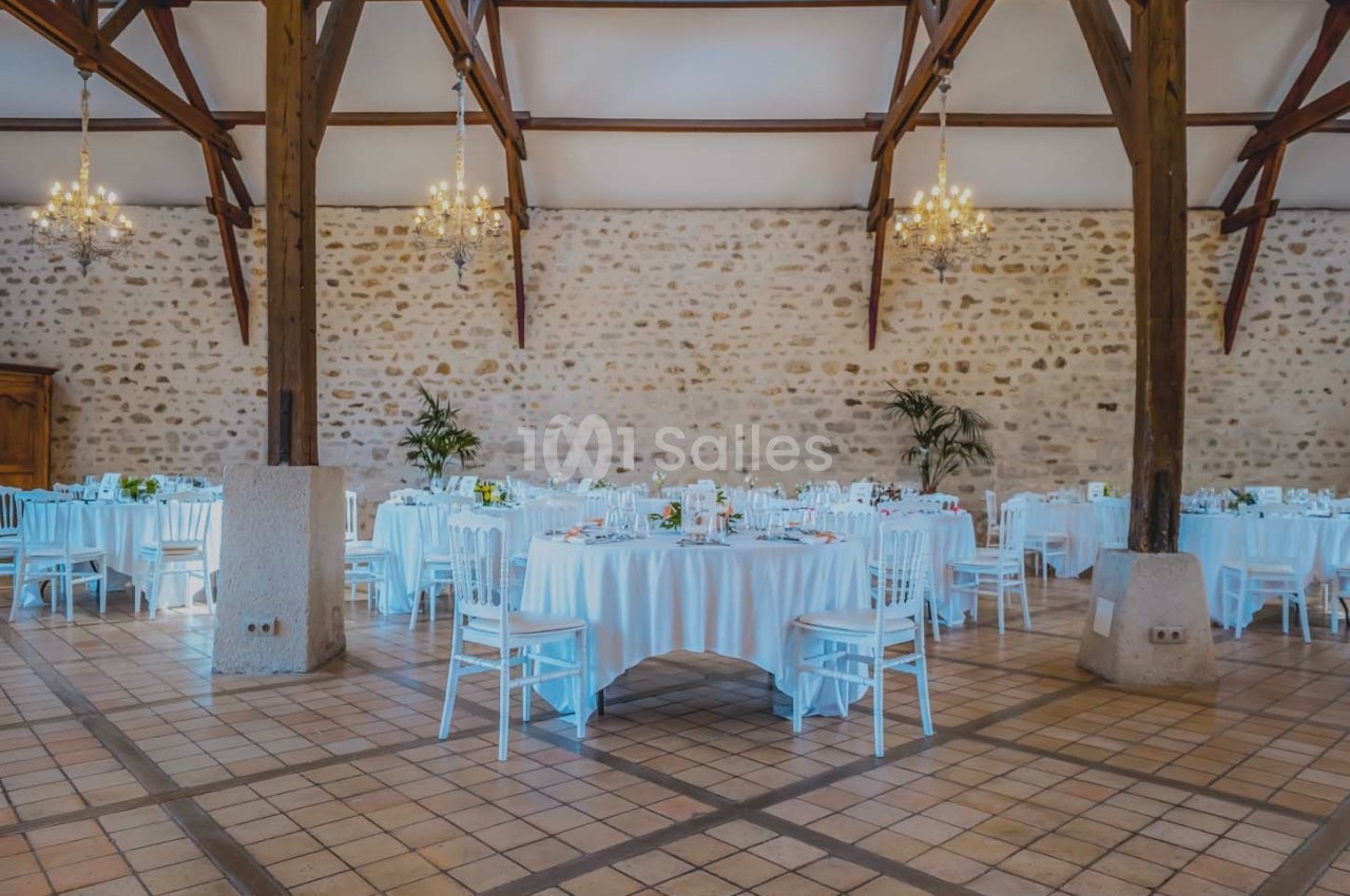 Salle de réception avec tables rondes dressées, chaises blanches, murs en pierre et lustres suspendus.