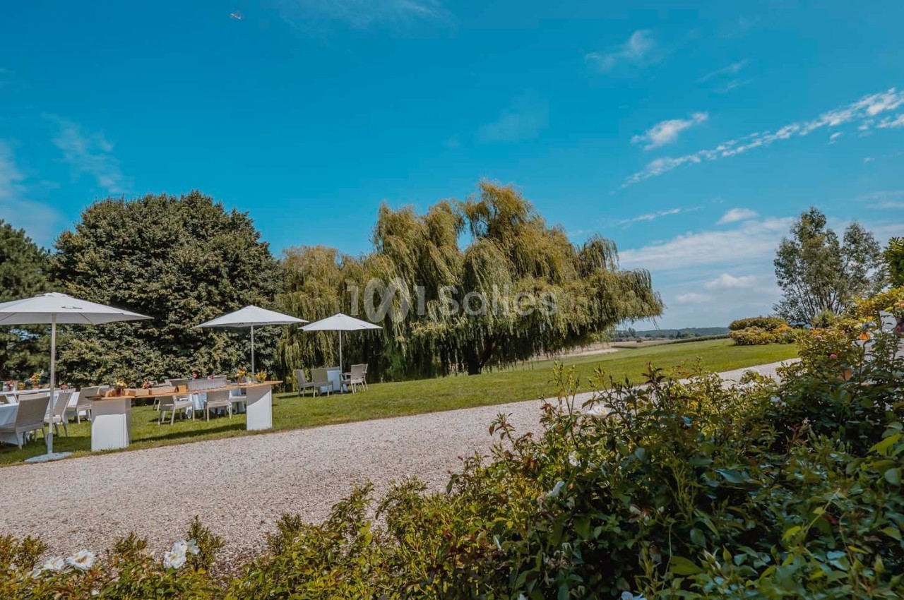 Espace extérieur avec tables et parasols près d'arbres et d'un chemin, sous un ciel bleu dégagé.
