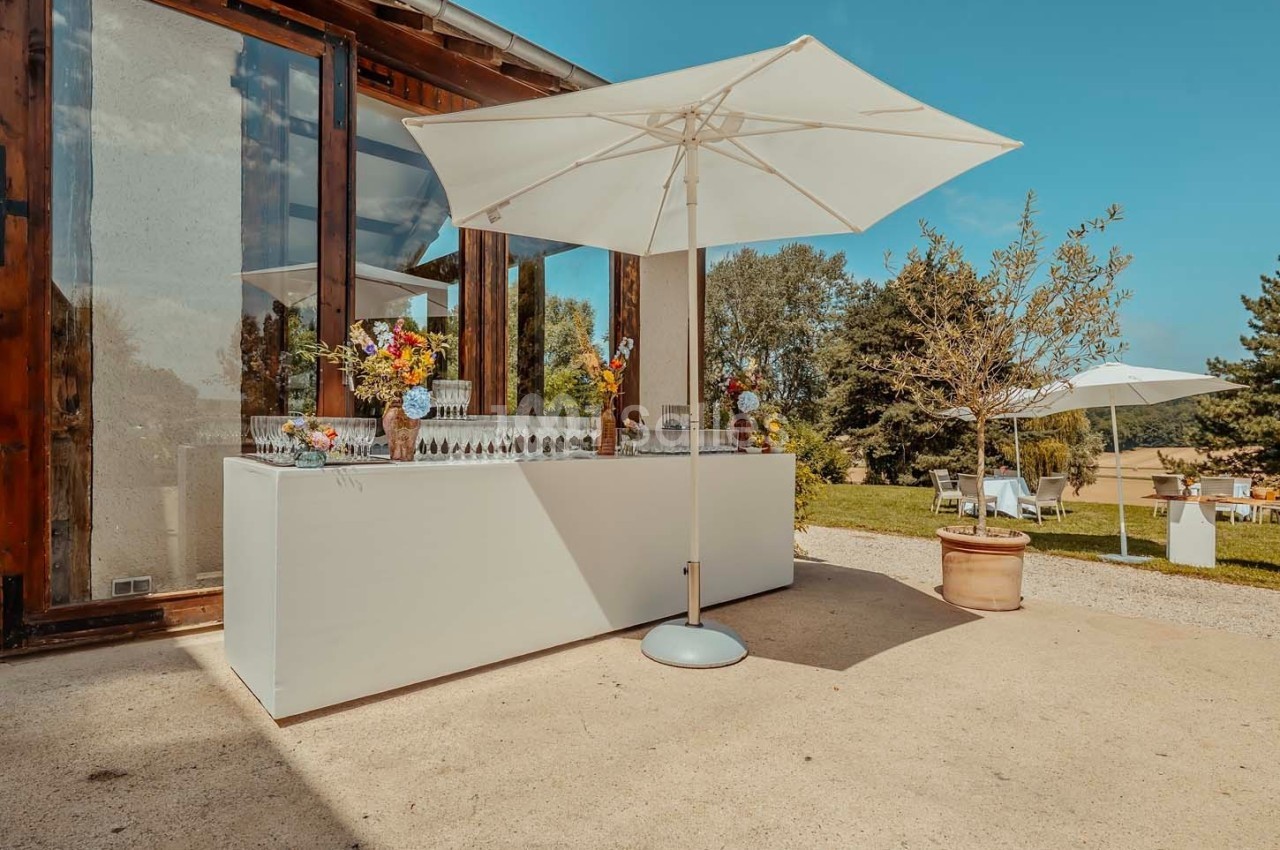 Table de réception extérieure avec verres, fleurs colorées et parasols, devant un bâtiment vitré et un jardin arboré.