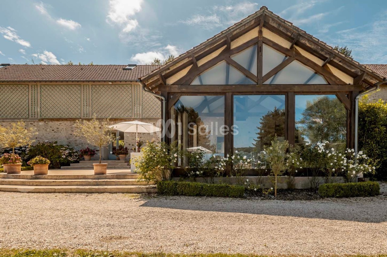 Façade d'un bâtiment avec une grande verrière, entourée de plantes et d'une terrasse avec parasol sous un ciel dégagé.