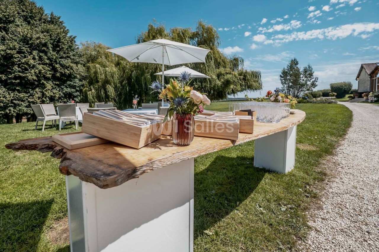 Table en bois brut avec des caisses en bois, des fleurs et des verres, installée dans un jardin sous des parasols.