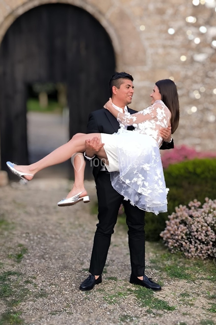 Un homme en costume noir porte une femme en robe blanche devant une porte en pierre dans un jardin fleuri.