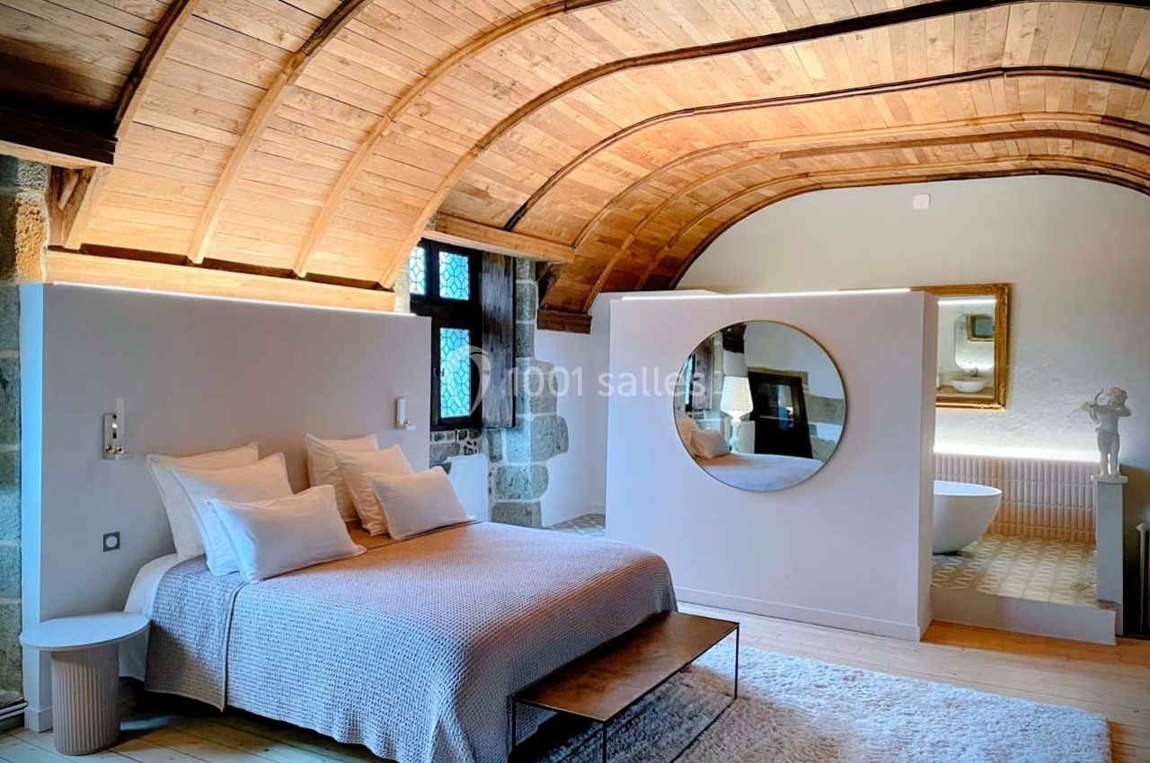 Chambre lumineuse avec plafond voûté en bois, lit double, miroir rond et salle de bain attenante visible.