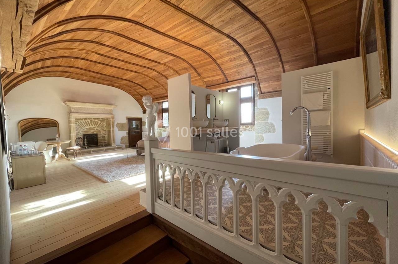 Chambre lumineuse avec plafond voûté en bois, espace salle de bain ouvert, cheminée et mobilier ancien.