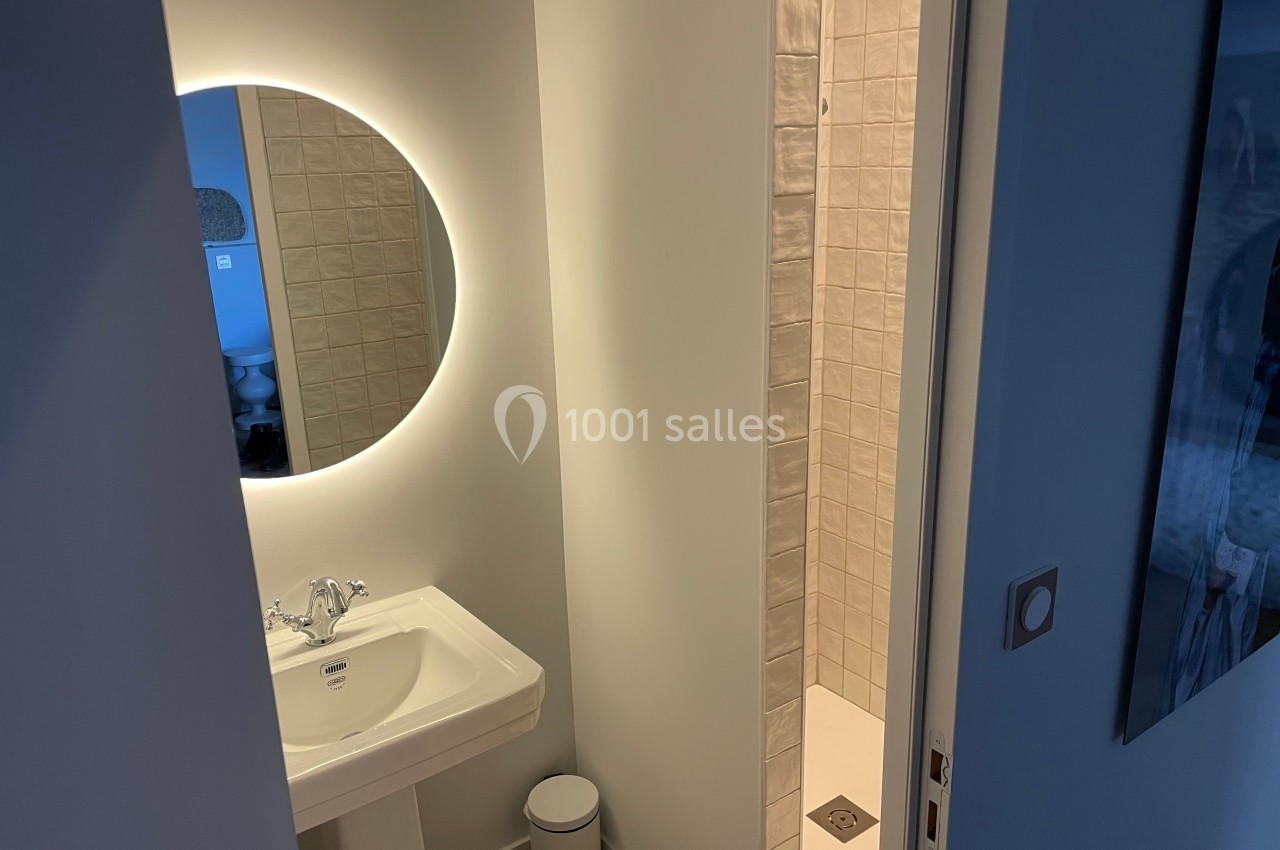 Salle de bain avec un lavabo, un miroir rond éclairé et une douche à carrelage clair visible en arrière-plan.