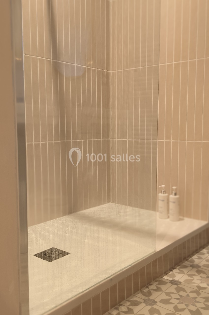 Douche moderne avec paroi vitrée, carrelage beige et sol à motifs géométriques, deux flacons posés au fond.
