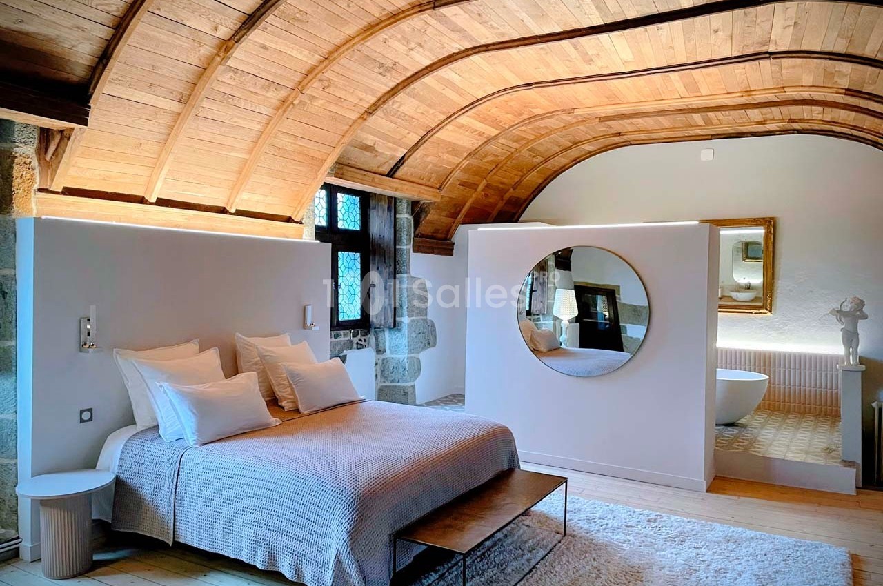 Chambre lumineuse avec plafond voûté en bois, lit double, miroir rond et salle de bain attenante visible.
