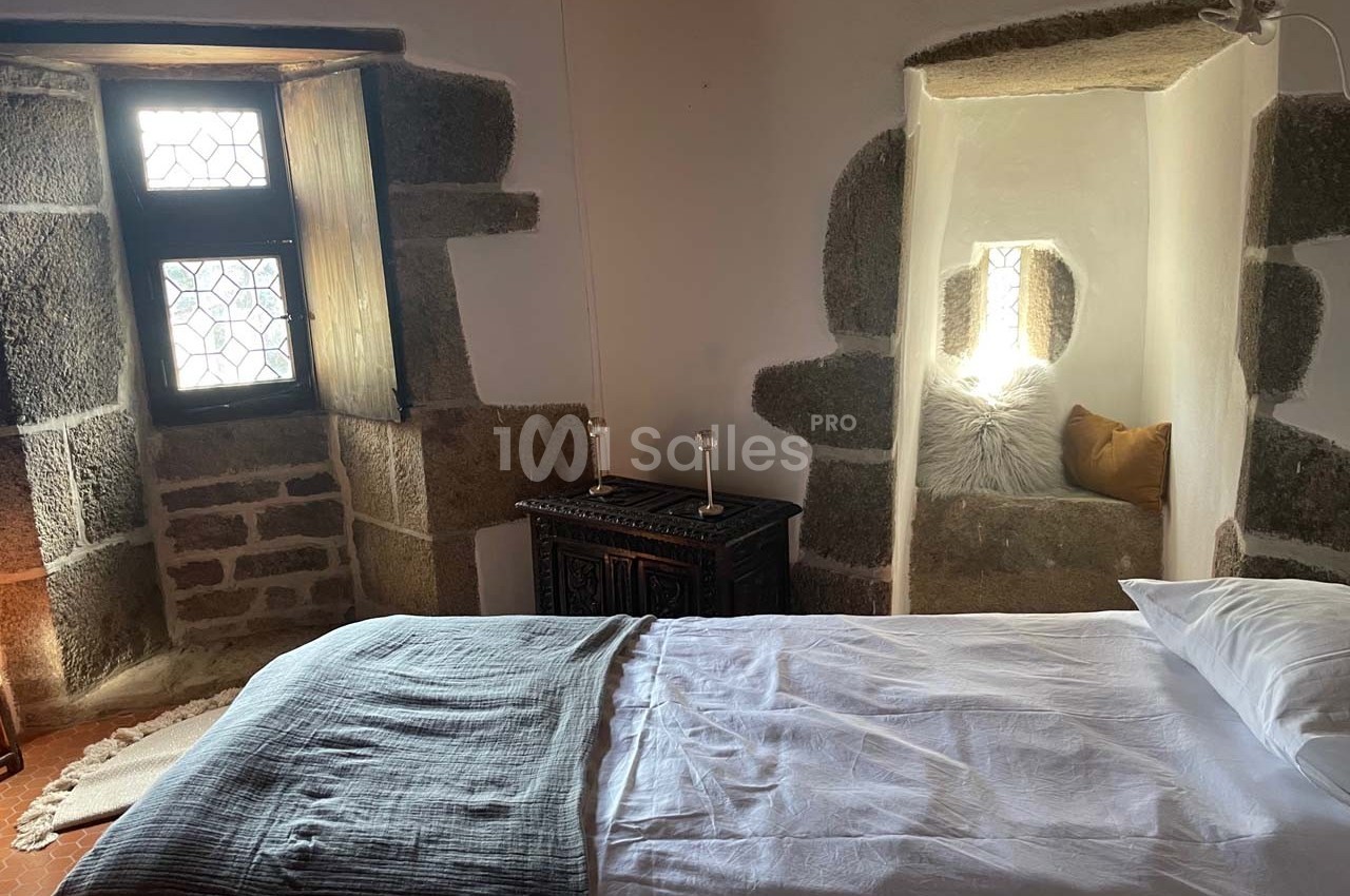 Chambre rustique avec lit simple, murs en pierre, fenêtre à vitraux et alcôve décorée de coussins.