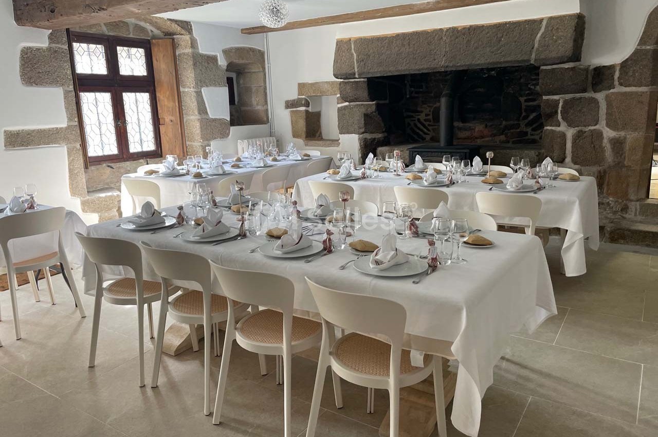 Salle de réception avec tables dressées, nappes blanches, vaisselle élégante et cheminée en pierre en arrière-plan.