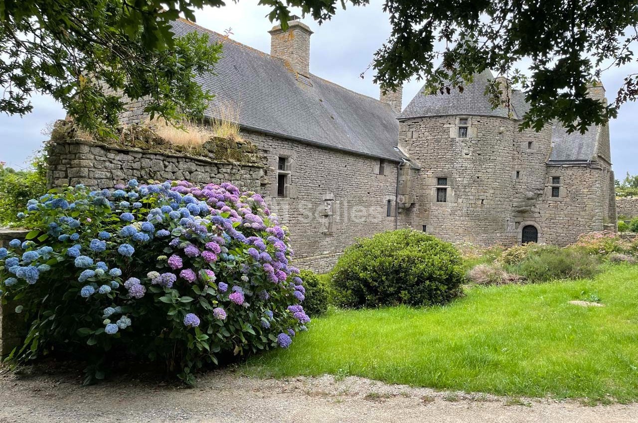 Vieille bâtisse en pierre entourée de verdure, avec des hortensias en fleurs au premier plan.