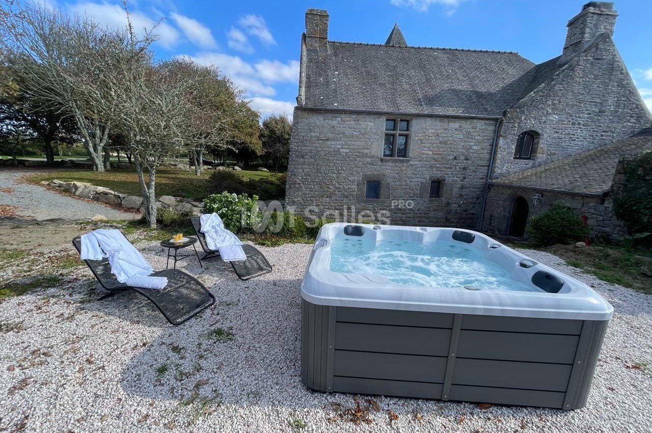Jacuzzi extérieur bouillonnant devant une maison en pierre, entouré de chaises longues et d'arbres sous un ciel dégagé.