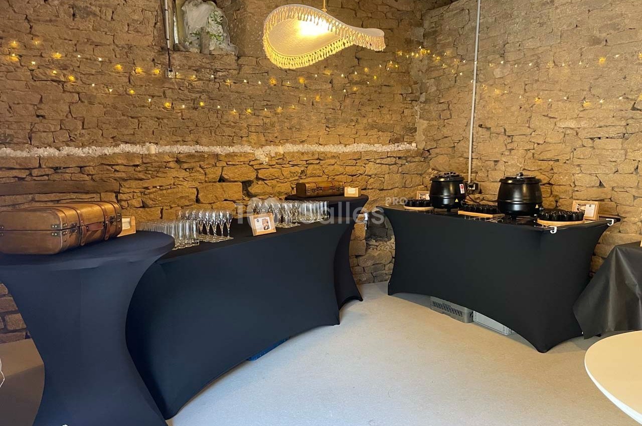 Buffet avec nappes noires, soupières, verres et décorations lumineuses dans une salle en pierre.