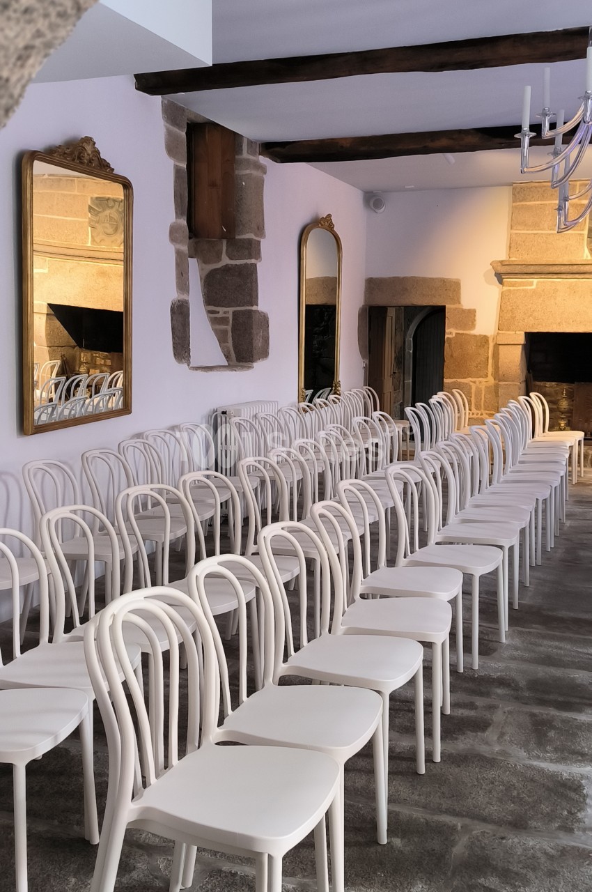 Salle en pierre aménagée avec des rangées de chaises blanches alignées, deux miroirs accrochés aux murs.