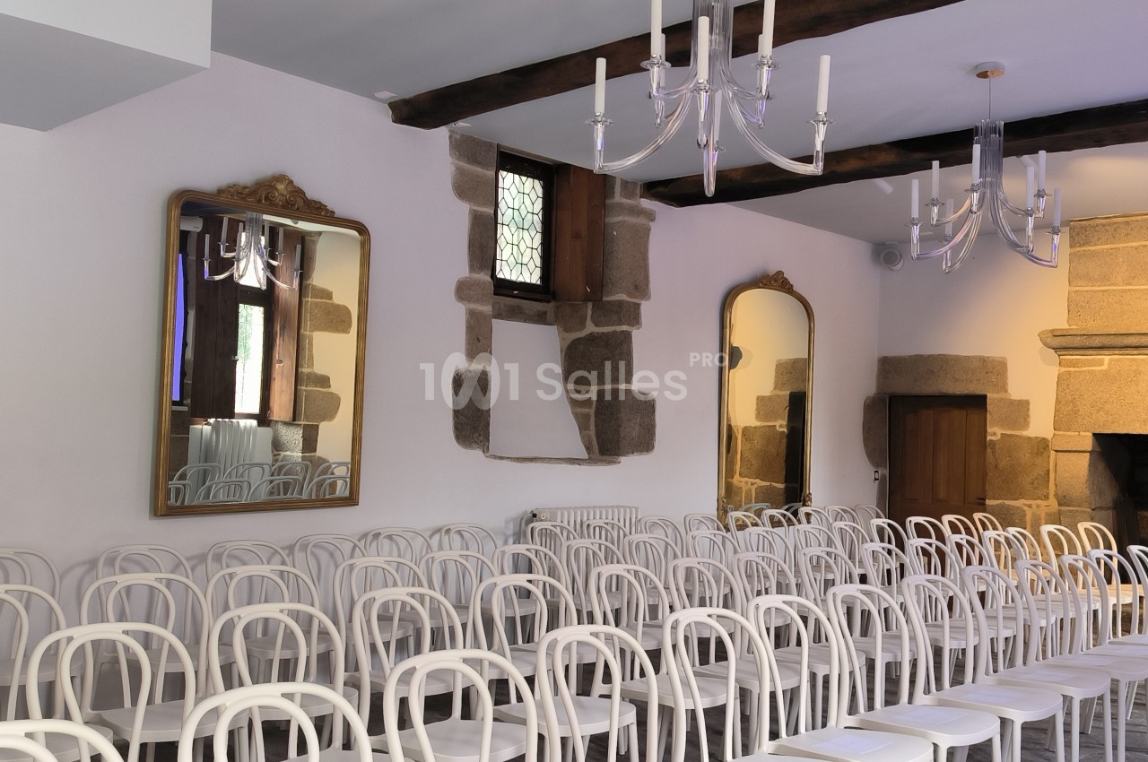Salle avec des chaises blanches alignées, murs en pierre, miroirs, lustres et une grande cheminée en arrière-plan.