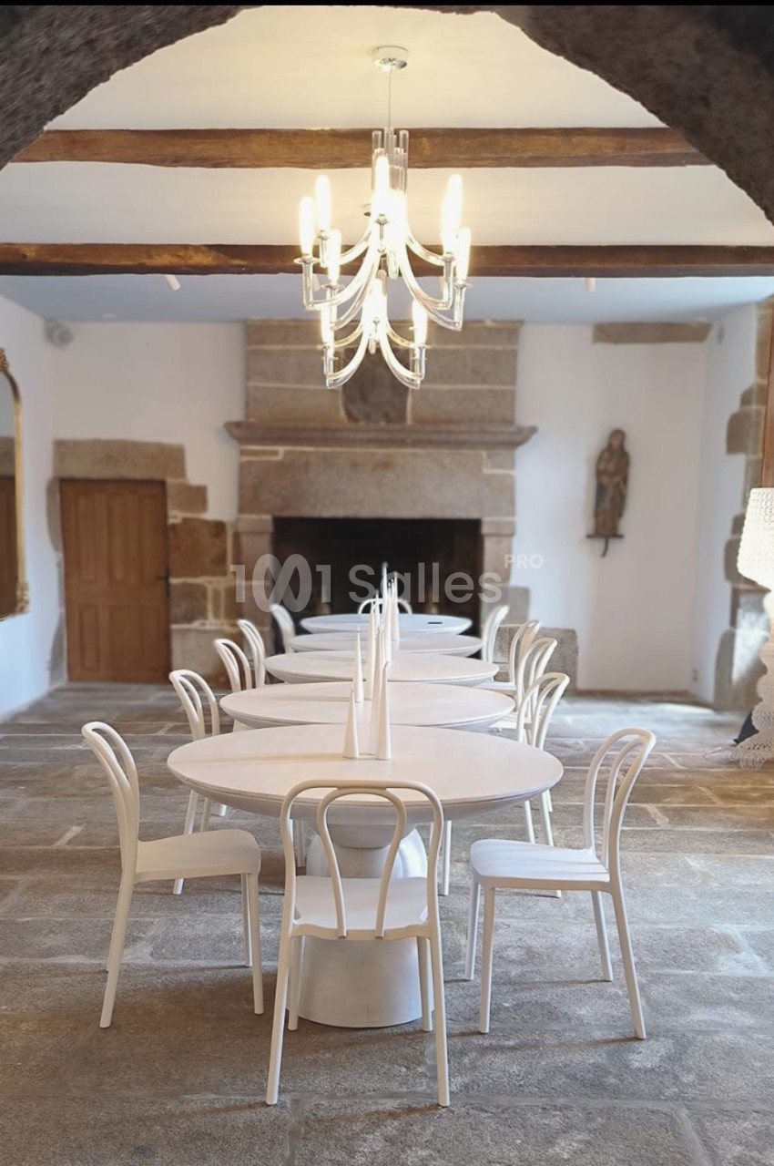 Salle lumineuse avec tables rondes blanches, chaises assorties, lustre moderne et cheminée en pierre.