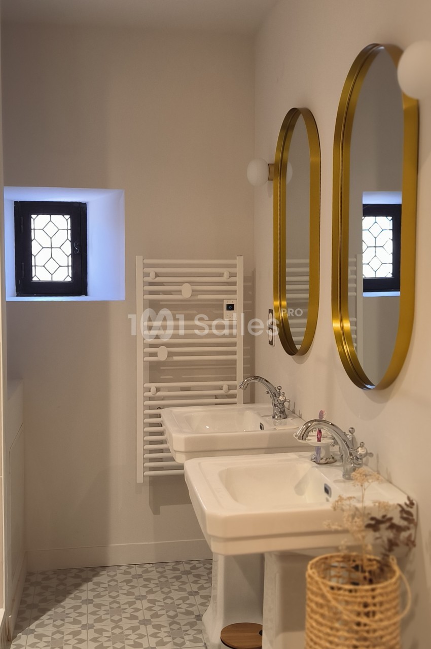 Salle de bain lumineuse avec deux lavabos, miroirs dorés, radiateur blanc et petite fenêtre à grille.