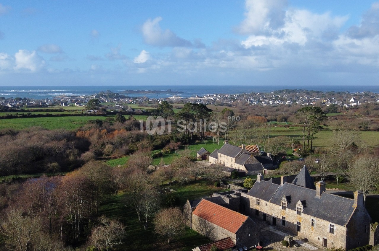 Vue aérienne d'un manoir en pierre entouré de champs et de végétation, avec un village et la mer à l'horizon.