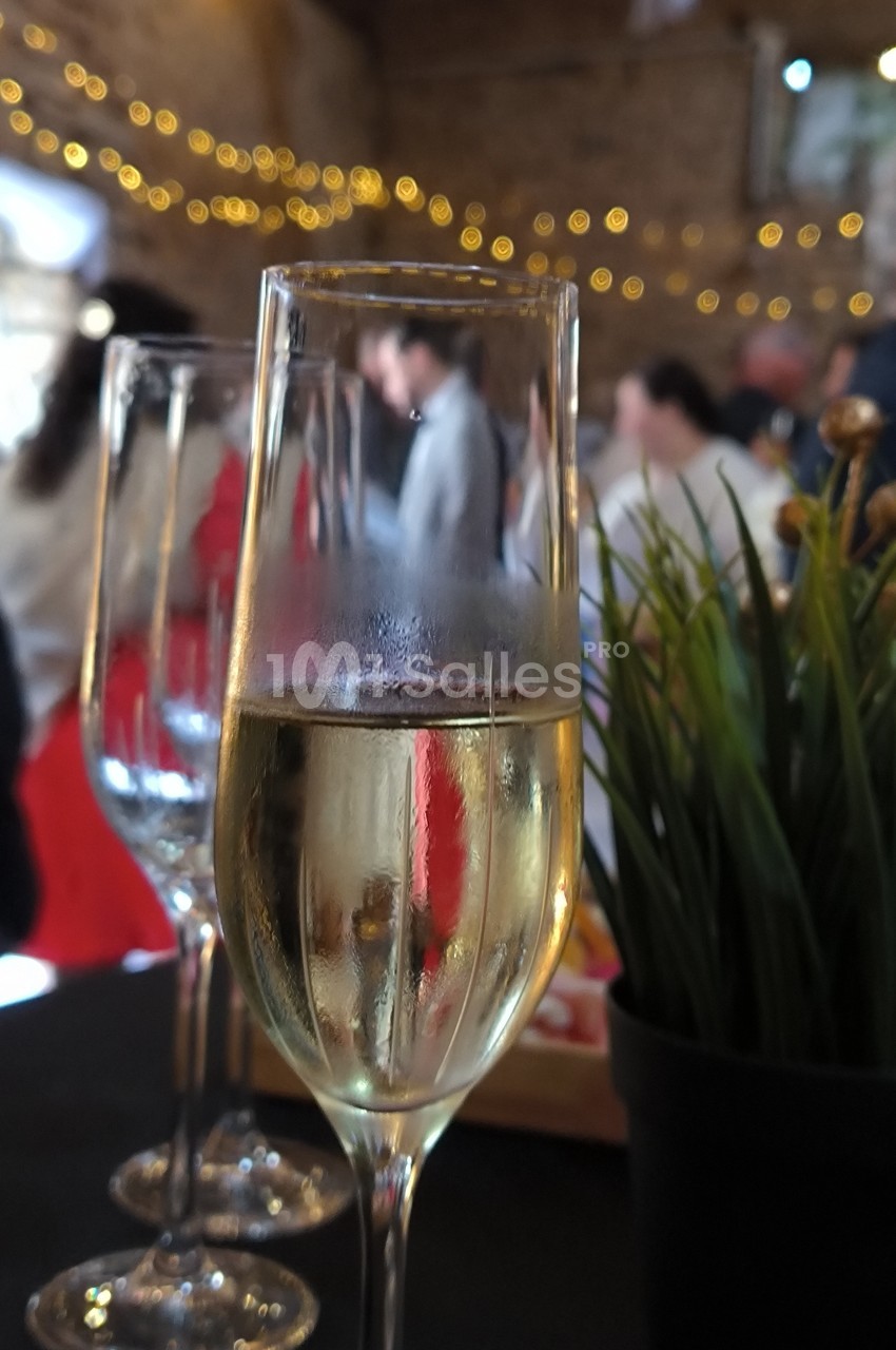 Verre de champagne sur une table, avec des invités flous en arrière-plan et des lumières décoratives.