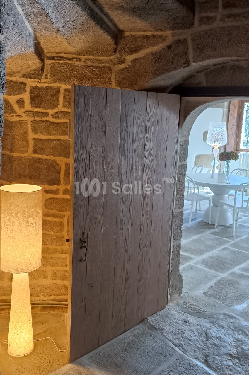 Porte en bois entrouverte dans un intérieur en pierre, laissant entrevoir une salle lumineuse avec des chaises blanches.